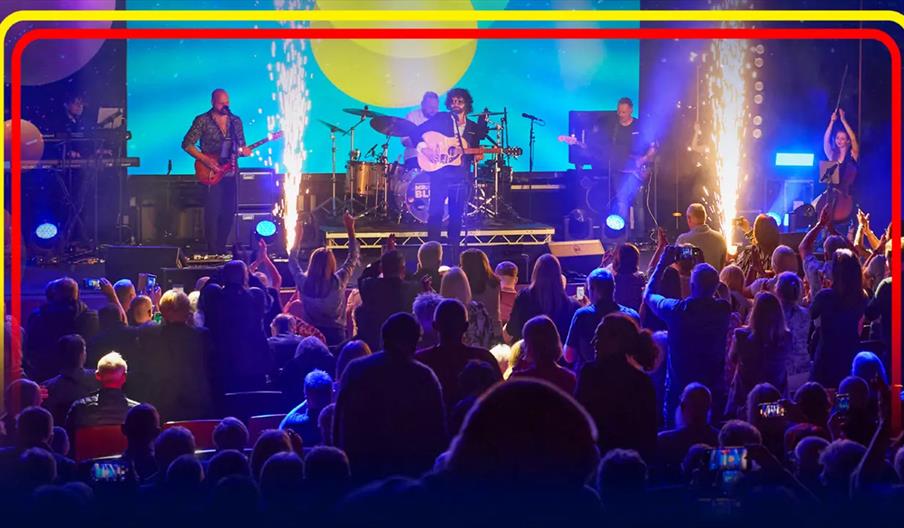 A band performs on stage with colourful lights and pyrotechnics as a large audience watches, some standing and taking photos, in front of a vibrant di
