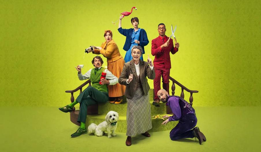 Six adults in colorful, eclectic outfits pose dramatically on a small green staircase against a bright green background. Props include an oversized pa