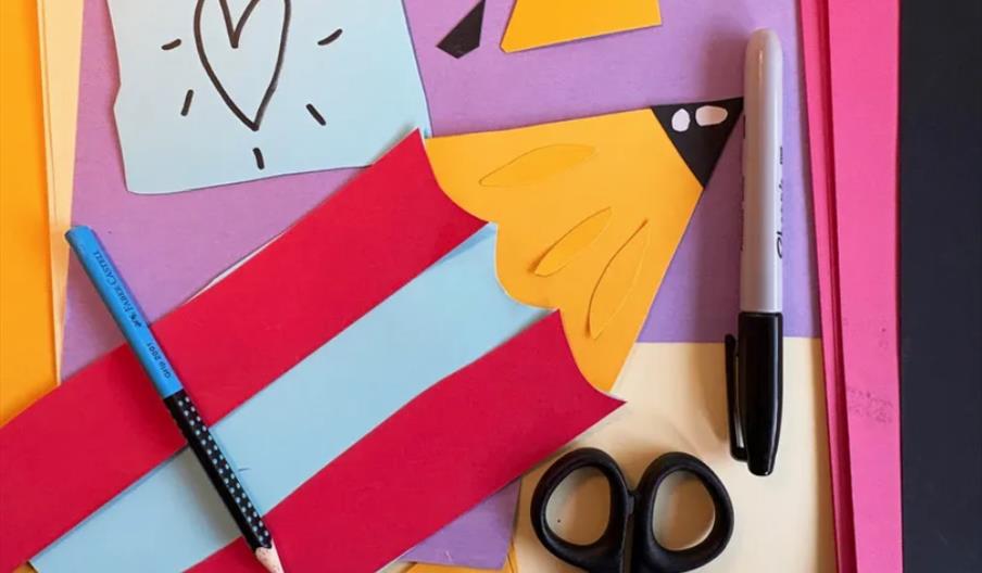 A collection of colourful construction paper cutouts, a blue pencil, a black marker, and a pair of black scissors are arranged on a flat surface; one