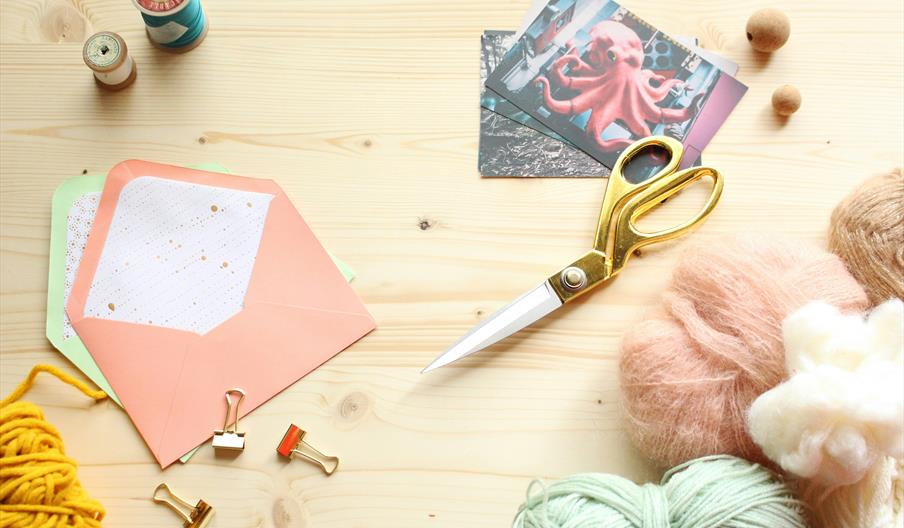 A flat lay of craft supplies on a light wooden surface, including golden scissors, colored yarn, spools of thread, pastel envelopes, binder clips, woo