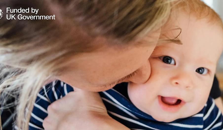 A close-up of an adult holding and kissing a smiling baby who is wearing a striped shirt, with the text "Funded by UK Government" in the top left corn