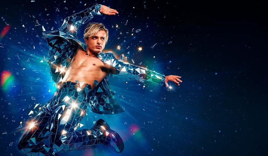 Nikita Kuzmin, wearing a shiny suit made of mirror-like glass shards, is leaping in the air against a dark, starry background as reflective fragments