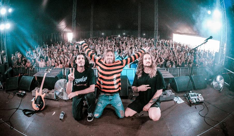 Three band members kneel at the front of a stage, smiling and making hand signs, with a large, enthusiastic crowd cheering behind them in an indoor co