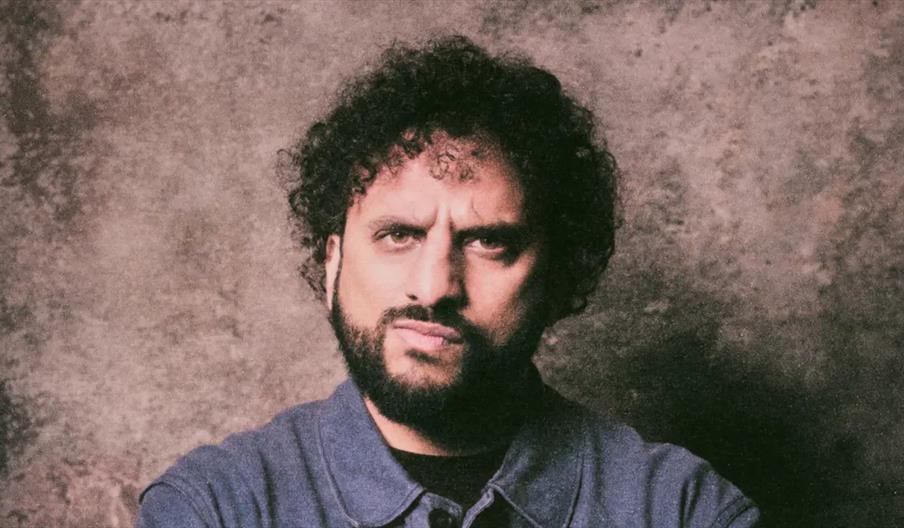 Nish Kumar with curly dark hair and a beard stands in front of a textured, neutral-colored backdrop, wearing a blue collared shirt and looking directl