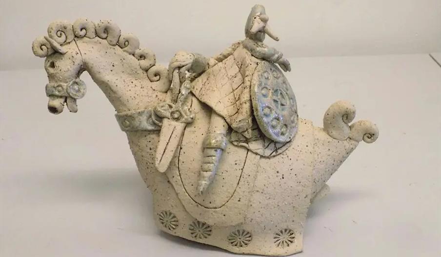 A ceramic sculpture of a stylized horse with curled details and a rider holding a shield and wearing armor, rendered in a rough, speckled white finish