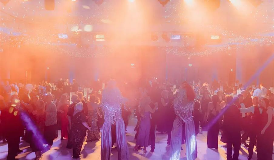 A large crowd of people in formal attire are dancing in a ballroom filled with fog and bright orange stage lights, with two performers on stilts in sp