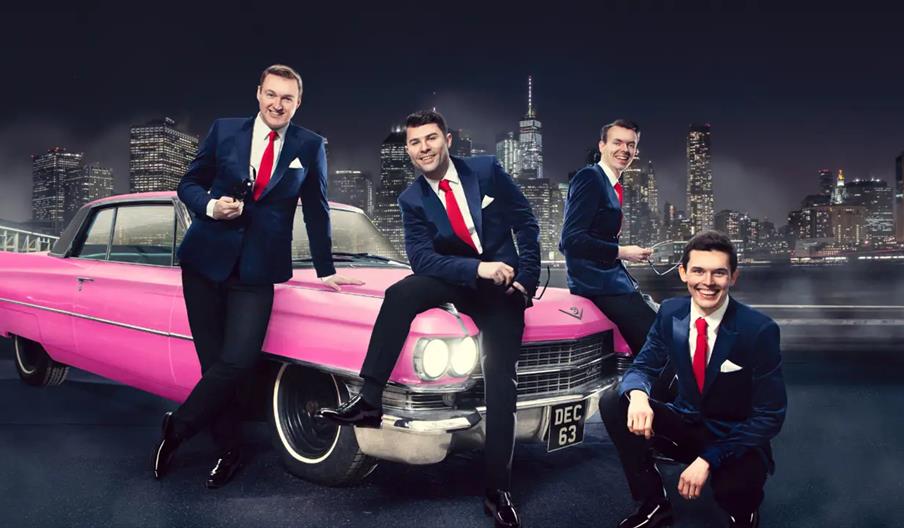 Four men wearing matching dark blue suits with red ties and white shirts pose in front of a bright pink vintage car at night, with a city skyline feat