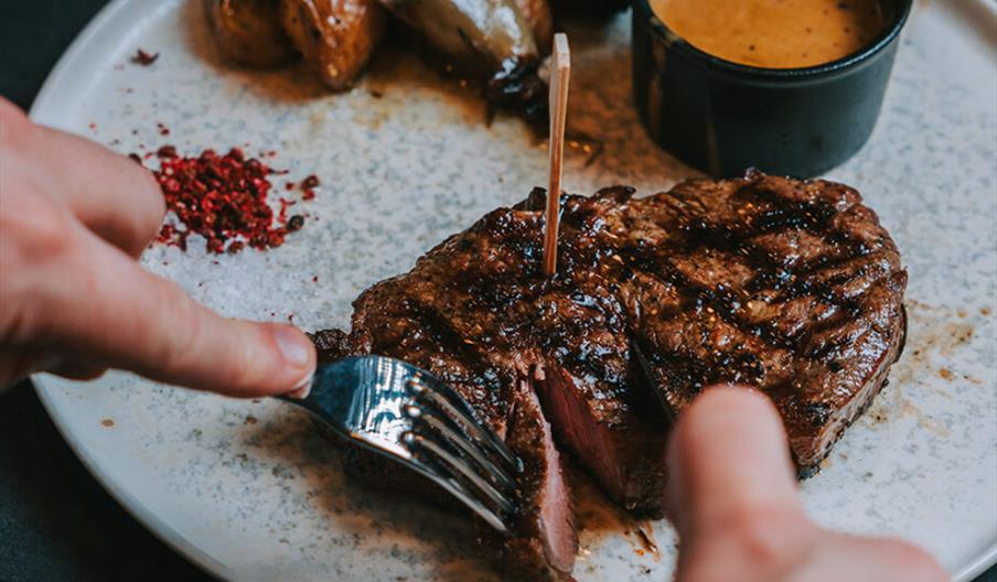 A portion of steak is being cut into on a plate with a side of potatoes and some peppercorn sauce.