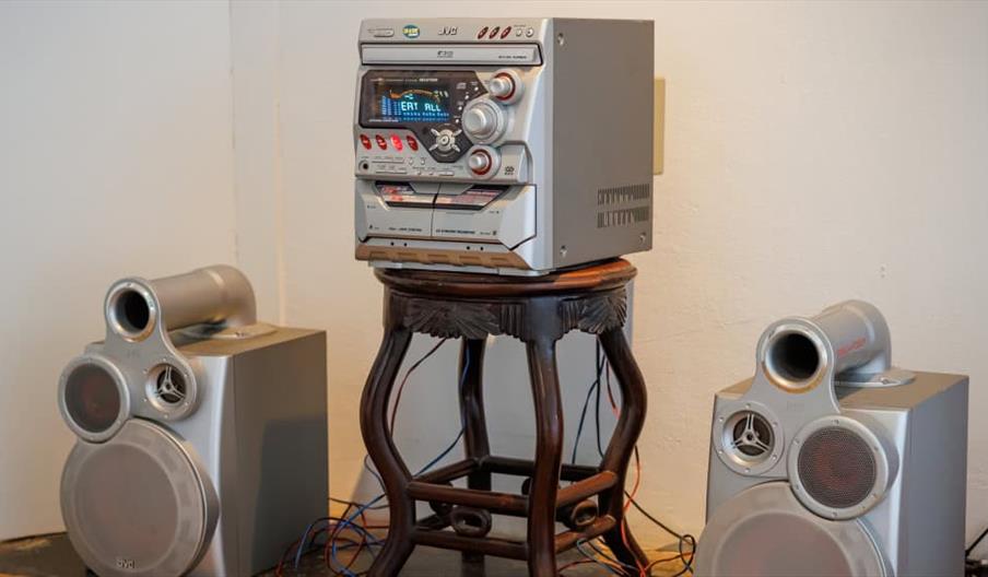 A silver JVC stereo system with dual cassette decks sits on a small wooden table, flanked by two large matching silver speakers on the floor in a corn