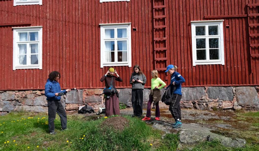 5 individuals stand in front of a red house with headphones.