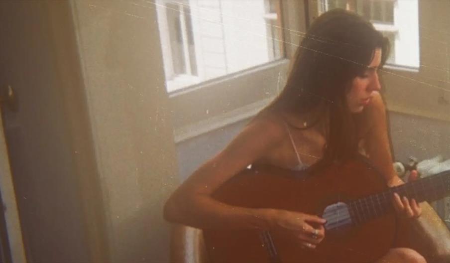 A young woman sits indoors by a window, playing an acoustic guitar and looking downward with a thoughtful expression. The image has a warm, vintage fi