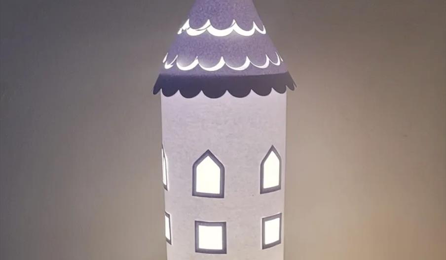 A cylindrical paper lantern designed to look like a fairytale tower, with arched windows cut out and a pointed, scalloped roof, softly glowing with li