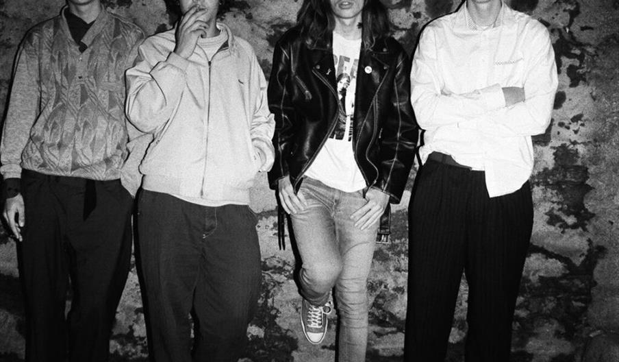 Black and white photo of four young men standing against a rough stone wall; one is smoking, and another wears a leather jacket and rests one foot aga