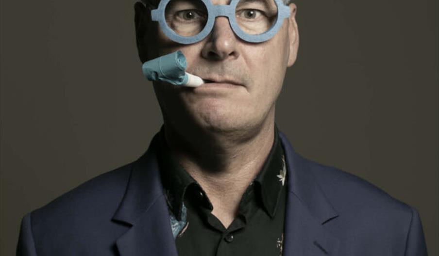 A man wearing a blue suit jacket and a dark floral shirt, with novelty blue glasses featuring white bunny ears and a party blower in his mouth, stands