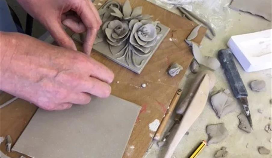 A person is shaping a clay tile decorated with sculpted flowers and leaves, surrounded by various pottery tools, scraps of clay, and a pencil on a wor