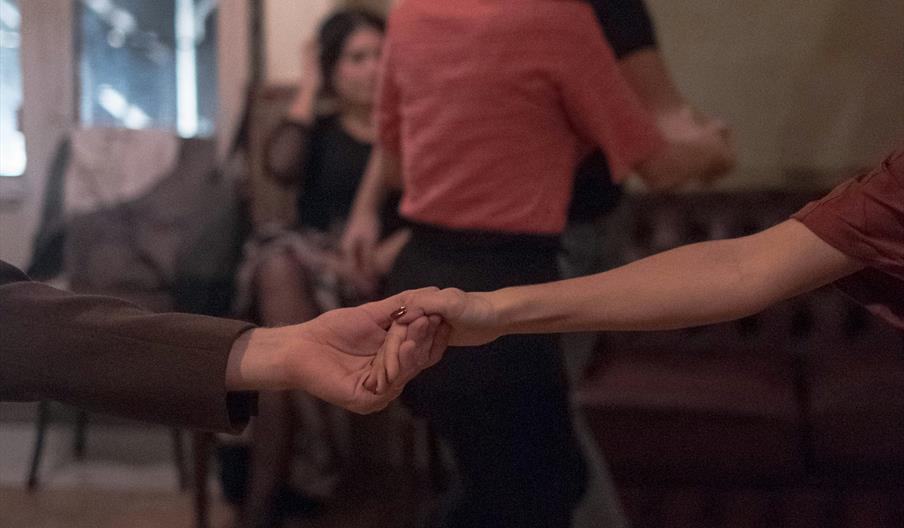 Two people are holding hands mid-dance in a dimly lit room, while another couple dances in the background and a woman sits on a chair watching.