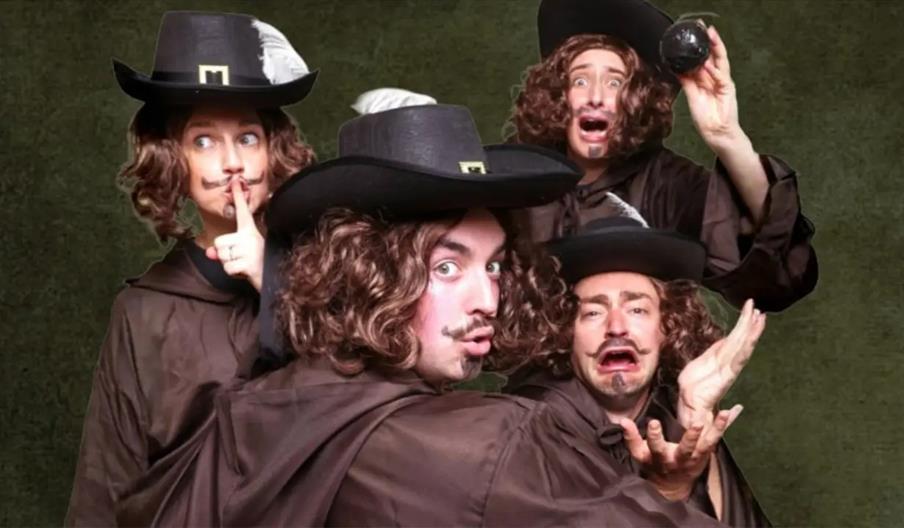Five people wearing curly brown wigs, fake mustaches, and black hats with white feathers pose together in dramatic and humorous expressions, dressed i
