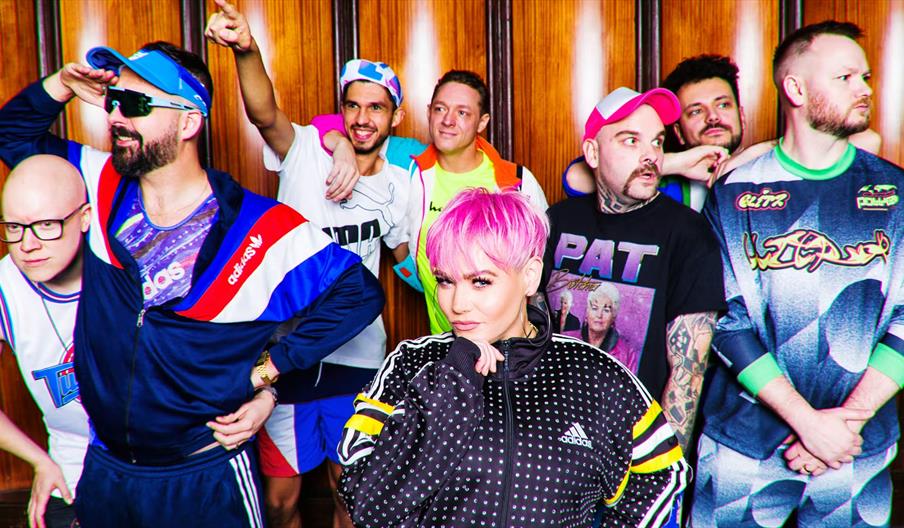 A group of seven people poses in front of a wooden wall, dressed in colorful, retro athletic clothing. The woman in the center with bright pink hair s