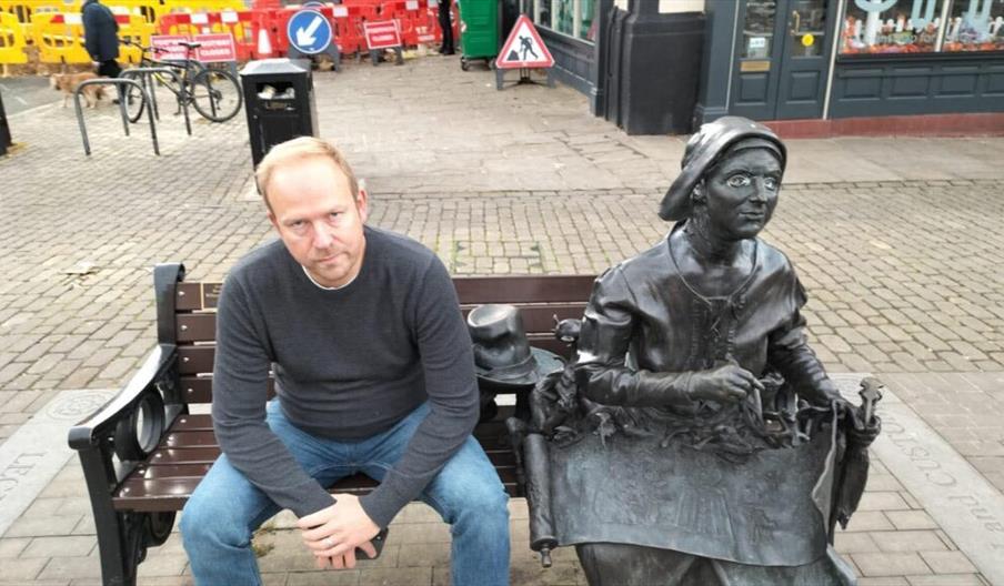 Comedian Rich Austin sat on a bench next to a statue of a woman.