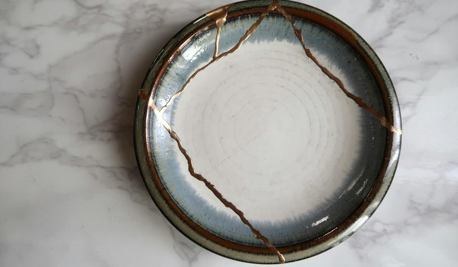 A ceramic plate repaired with the kintsugi technique, featuring visible gold lines where cracks were mended, placed on a light marble surface.