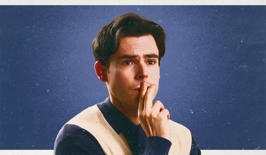 A young man with dark hair is touching his lips with his fingers and looking thoughtful, standing in front of a blue background.