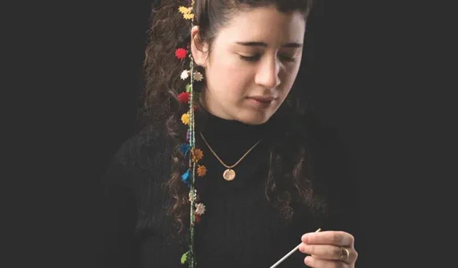 A woman with curly hair and a ponytail decorated with colorful yarn flowers looks down as she works with a crochet hook. She is wearing a black top an
