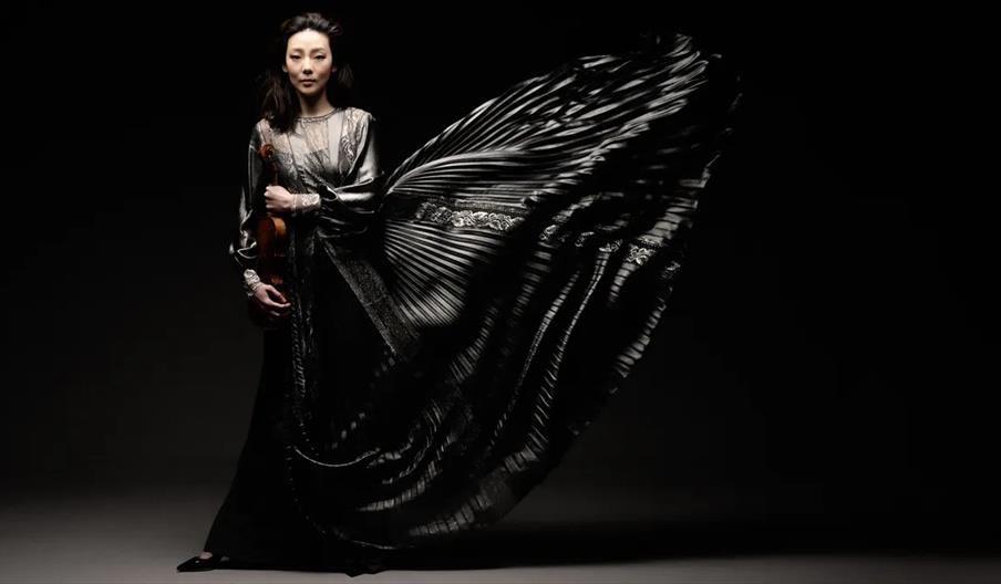 A woman in a dramatic, flowing black gown stands against a dark background, holding a violin; the fabric of her dress is gracefully fanned out to one