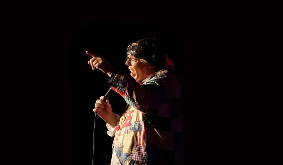 Roy Chubby Brown wearing his colourful patchwork jacket and aviator goggles holds a microphone and points while performing on a dark stage, illuminate