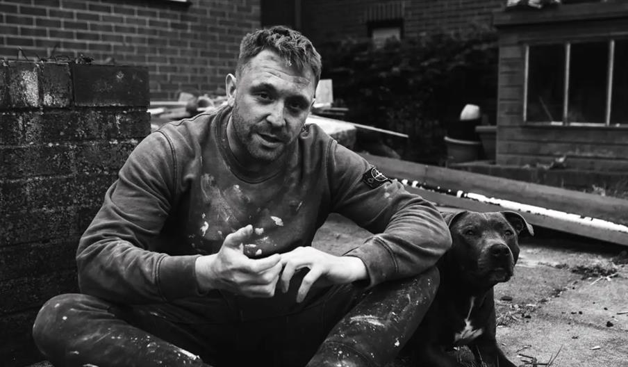 A man with a dirty, paint-splattered sweatshirt sits on the ground outdoors next to a black dog. The background shows brick walls, construction materi