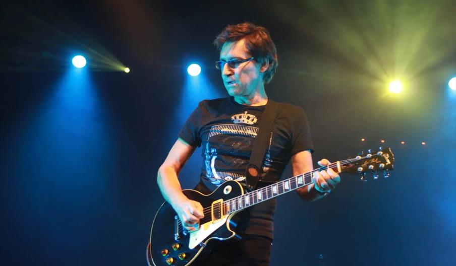A musician wearing glasses and a black t-shirt plays an electric guitar on stage, illuminated by colorful blue and yellow stage lights.