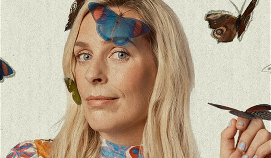 Sara Pascoe is wearing a colorful, floral-patterned blouse is surrounded by butterflies, with one blue butterfly resting on her forehead and another o