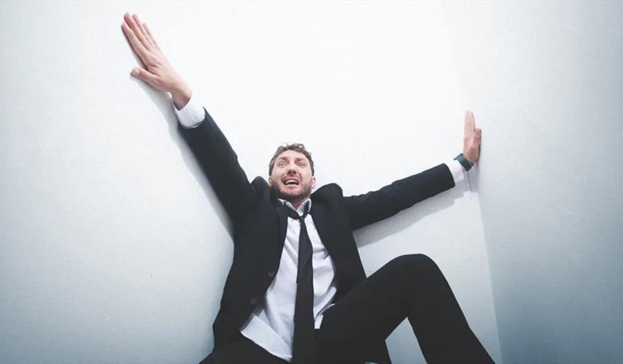 Seann Walsh in a business suit sits on the floor in a tight corner with his arms stretched out, looking anxious or stressed.