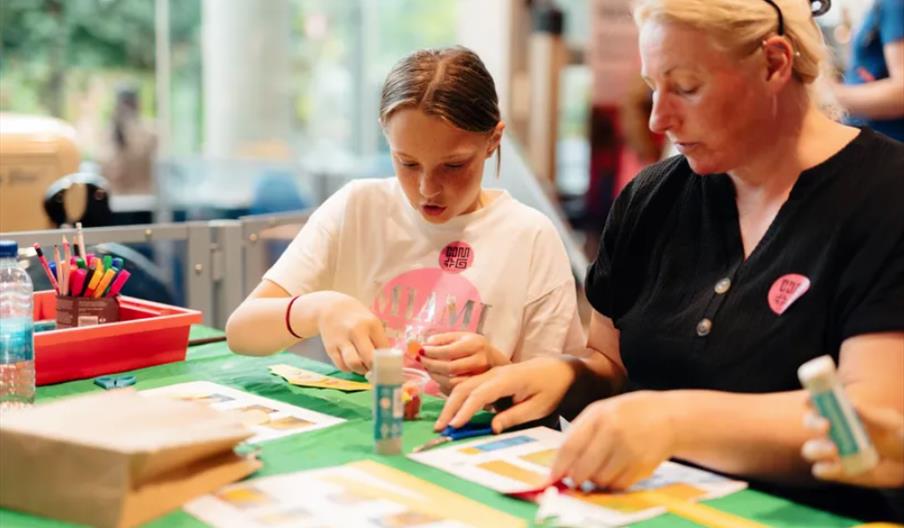 A girl and a woman sit at a table covered with craft supplies, including colored pens, glue sticks, and paper, as they work on an arts and crafts proj