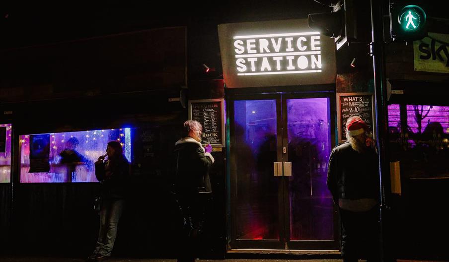 Nighttime street view of a venue called "Service Station" with a brightly lit sign above the entrance, two people standing outside, and purple lightin