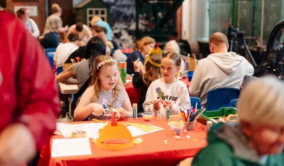 Two young girls sit at a red table covered with art supplies, crafting and talking, while other children and adults are busy with similar activities i