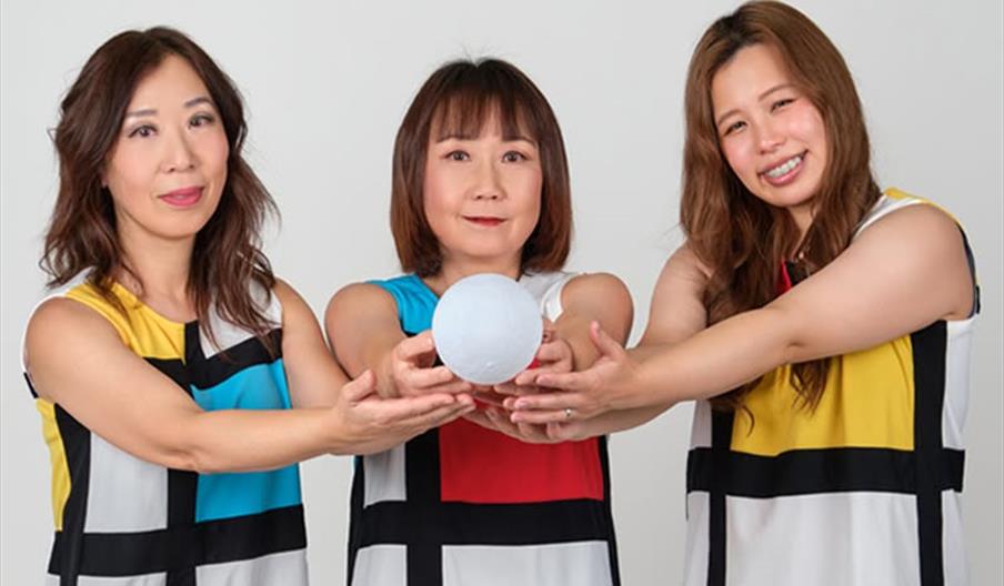 Three women wearing color-block patterned dresses stand side by side, smiling and holding a white ball together in front of them against a plain backg
