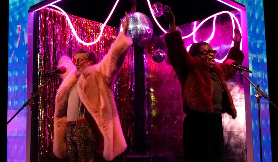 Two performers wearing colorful faux fur coats stand on stage with microphones, raising their arms in celebration. The stage is decorated with bright