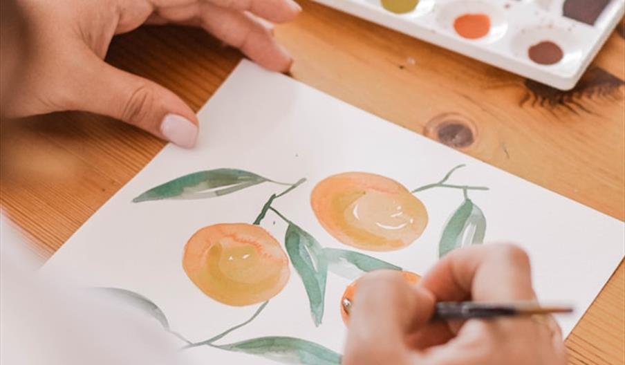 A person is painting a watercolor picture of oranges with green leaves on white paper, using a brush and a palette of watercolor paints on a wooden ta