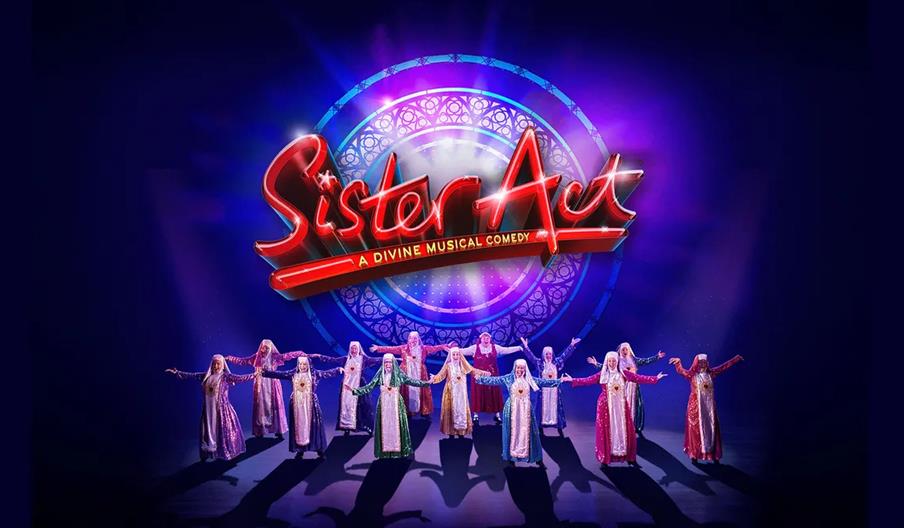 A group of performers dressed as nuns in brightly colored habits stand in a line on stage with their arms outstretched, beneath a large illuminated "S