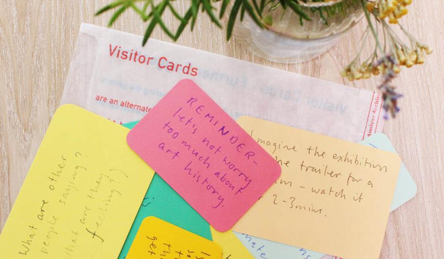 A tabletop with colorful handwritten visitor cards, including a pink card that says "REMINDER – let's not worry too much about art history," and a sma