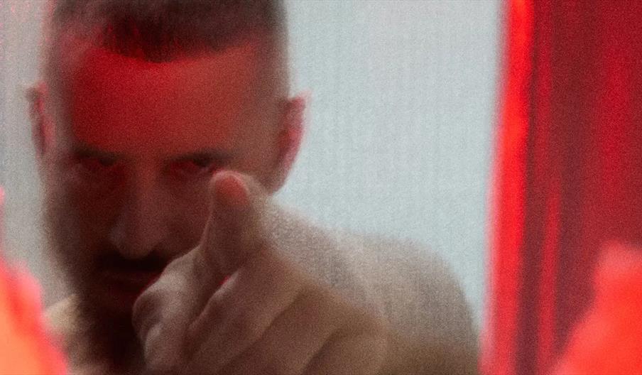 A blurry, close-up image of a person pointing directly at the camera, with red lighting partially illuminating their face and hand. The background and