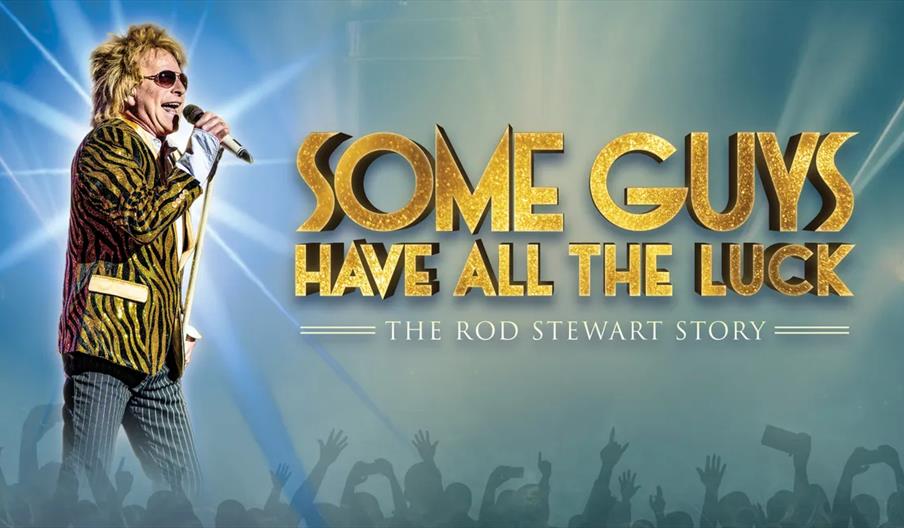 A performer with spiky blonde hair, sunglasses, and a flashy gold-and-black jacket sings into a microphone on stage. The background features bright st