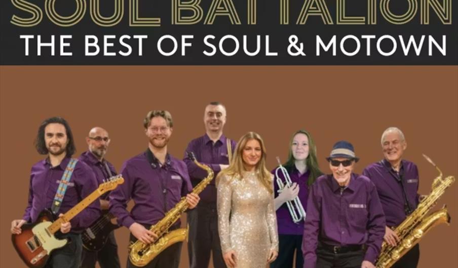 A group photo of eight band members, most wearing matching purple shirts, with various musical instruments including guitars, saxophones, and a trumpe