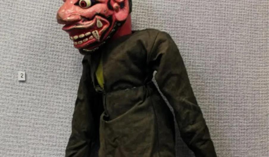 A traditional puppet with a large, exaggerated red face, wide eyes, sharp teeth, and black hair, wearing a dark shirt and patterned skirt, is displaye