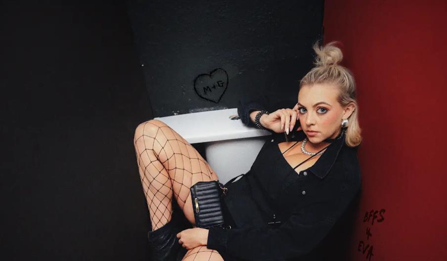 A young woman with blonde hair in a messy bun, wearing a black outfit, fishnet stockings, and boots, lounges in a bathtub against black and red walls