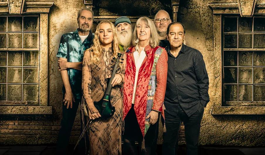 Six adults stand closely together in front of a textured wall with old windows; one woman in front holds a violin.