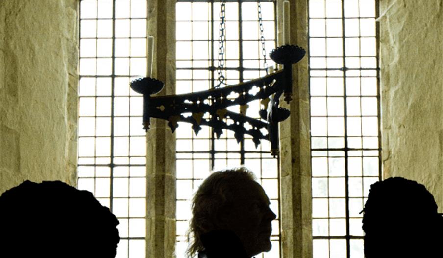 Three people are silhouetted in front of tall, arched windows and a hanging chandelier inside a church or cathedral, with light streaming through the