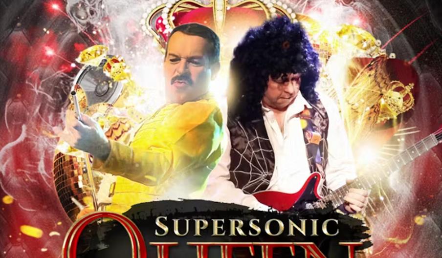 Two performers on stage dressed as Freddie Mercury and Brian May from Queen, with one singing into a microphone and the other playing electric guitar.