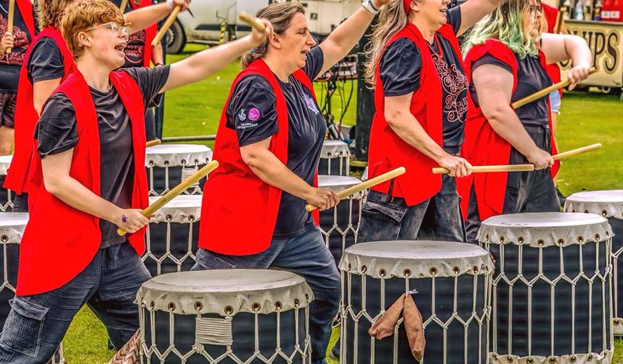 A group of people in matching red vests enthusiastically play large barrel drums outdoors, raising drumsticks in synchronized motion, with a festival