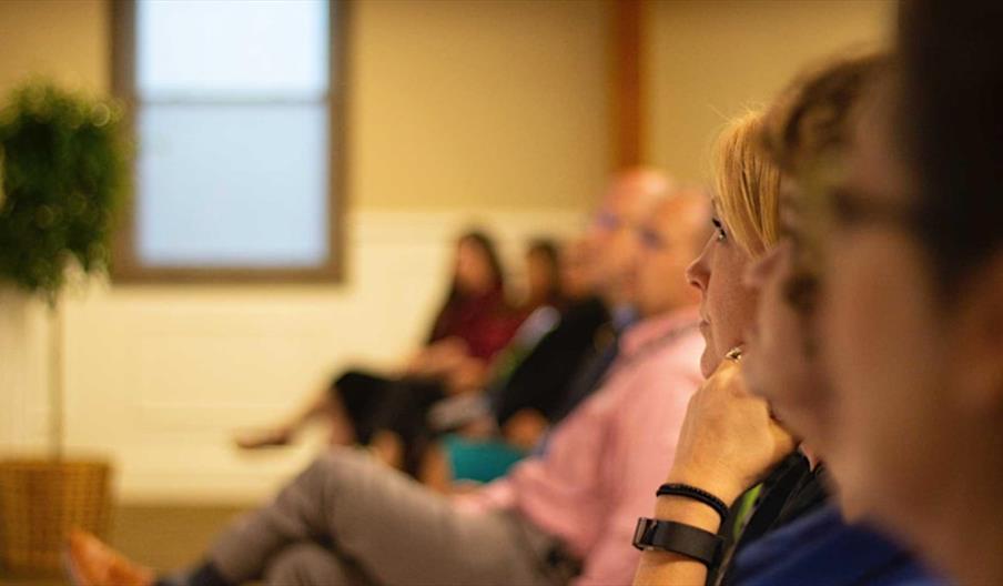 A group of people seated indoors in a classroom or seminar setting, all facing forward and appearing attentive, with the image focused on the side pro